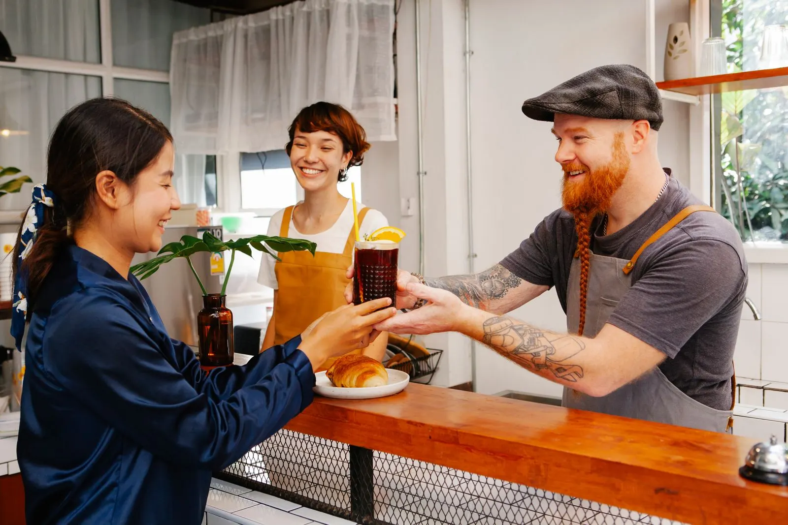 Χαμογελαστός barista με γένια και καπέλο δίνει έναν κρύο καφέ πάνω από ξύλινο πάγκο σε μια νεαρή πελάτισσα, σε φωτεινή καφετέρια